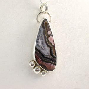 May include: A silver pendant with a teardrop-shaped agate stone. The stone has a swirling pattern of brown, black, white, and pink colors. The pendant has three silver beads at the bottom.