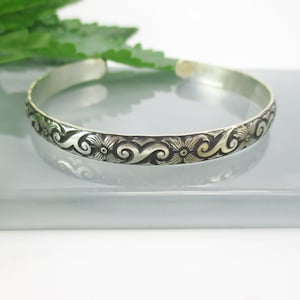 May include: A silver bangle bracelet with an intricate floral design. The design features swirls and flowers.