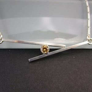 May include: A silver necklace with a geometric design featuring two intersecting bars and a small, round, yellow gemstone.