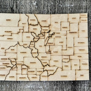 May include: A wooden map of Colorado with the names of each county labeled. The map is divided into 64 counties.