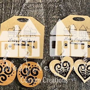 Puede incluir: Dos pares de pendientes presentados en cartulina marrón. Un par presenta pendientes redondos de madera con un diseño de espiral negro. El otro par tiene pendientes de madera en forma de corazón con un diseño de filigrana negro. Los pendientes cuelgan de ganchos plateados.