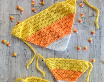 May include: A handmade crochet bikini set in yellow, orange, and white, resembling candy corn. The top and bottom pieces are triangular, with long ties. Candy corn pieces are scattered around the set on a wooden surface. A festive, unique swimwear option.