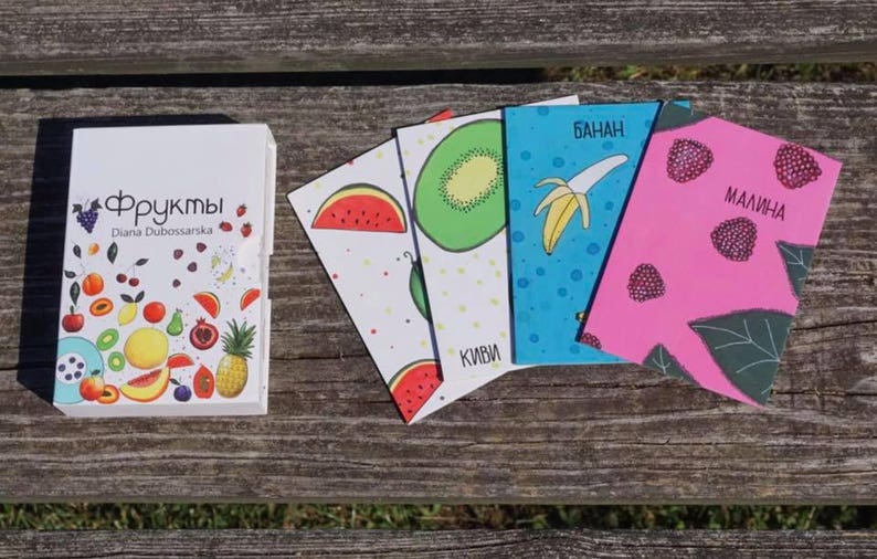 May include: A set of five colorful flashcards featuring different fruits. The cards are labeled with the Russian words for "watermelon", "kiwi", "banana", and "raspberry". The cards are arranged on a wooden surface.