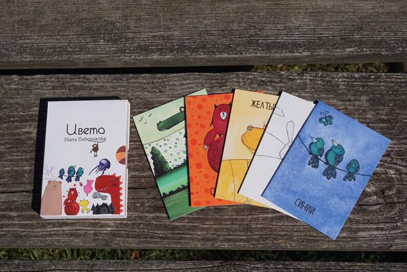 May include: A book titled "Ubema" by Diana Dubassenskaya, with colorful illustrations of animals. The book is next to five cards with illustrations of animals and the words "zheltyi" and "siniy" on them.