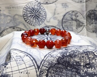 May include: A red carnelian gemstone bracelet with a silver magnetic clasp. The bracelet is made of round beads and is lying on a white fabric with a map pattern.
