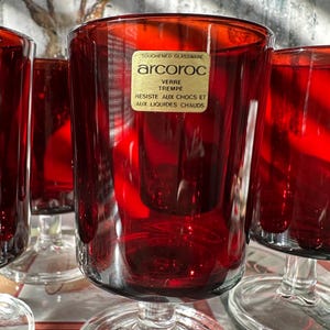 May include: A set of red Arcoroc stemmed glasses. The glasses have a clear base and a deep red bowl. The label on the glass reads "TOUGHENED GLASSWARE, Arcoroc, VERRE TREMPE, RESISTE AUX CHOCS ET AUX LIQUIDES CHAUDS."