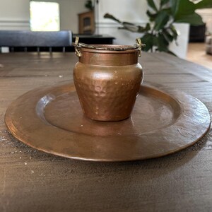 May include: A copper-colored, hammered metal pot with a lid and small handles, sitting on a matching copper plate. The pot and plate are on a wooden table. The pot has a textured surface and a slightly rounded shape.