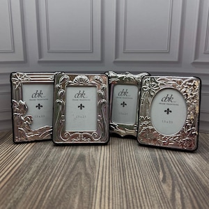 May include: Four silver-toned metal picture frames with intricate embossed floral and scrollwork designs. Each frame contains a white insert printed with the text cbk FRAME TRADITIONS and 1.5 x 2.5 inches. The frames feature rectangular and oval openings.