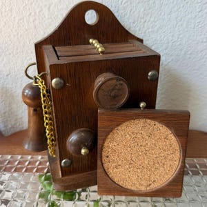 May include: A dark brown wooden coaster holder with a wall-mounting hole at the top. It features a gold-colored metal chain attached to a small wooden handle, brass-toned hardware, and a square wooden coaster with a circular cork center insert.