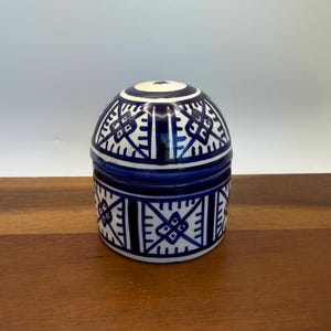 May include: A blue and white ceramic jar with a lid. The jar has a rounded shape with geometric patterns in dark blue against a white background. The jar is sitting on a wooden surface.