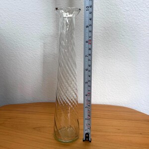 Pu&ograve; includere: Un vaso di vetro trasparente con un design a spirale, alto circa 25 cm. Il vaso ha una forma sottile e allungata con una parte superiore svasata. Il vaso &egrave; appoggiato su una superficie di legno.