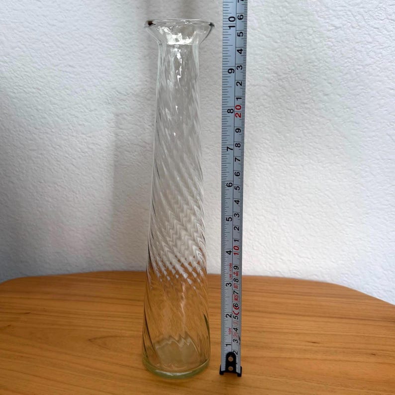 Pu&ograve; includere: Un vaso di vetro trasparente con un design a spirale, alto circa 25 cm. Il vaso ha una forma sottile e allungata con una parte superiore svasata. Il vaso &egrave; appoggiato su una superficie di legno.
