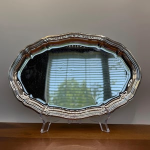 May include: A silver-colored, oval serving tray with a decorative, scalloped edge. The tray is displayed on a clear acrylic stand, reflecting the light and surrounding environment. The tray's surface is highly reflective.