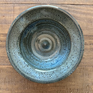 May include: A handmade ceramic bowl with a speckled blue glaze. The bowl has a wide rim and a shallow interior, with a spiral pattern in the center. The bowl is approximately 10 inches in diameter.