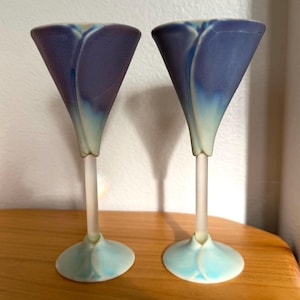 May include: Two decorative martini glasses with a unique design. The glasses feature a cone-shaped bowl in shades of purple and blue, transitioning to a frosted stem and a light blue base. The glasses are placed on a wooden surface.
