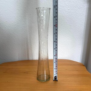 Pu&ograve; includere: Un vaso di vetro trasparente con un design a spirale attorcigliata. Il vaso &egrave; alto circa 25 cm. Ha una base svasata e un'apertura leggermente pi&ugrave; ampia nella parte superiore. Il vaso &egrave; appoggiato su una superficie di legno.