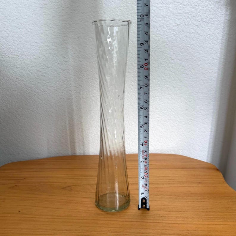 Pu&ograve; includere: Un vaso di vetro trasparente con un design a spirale attorcigliata. Il vaso &egrave; alto circa 25 cm. Ha una base svasata e un'apertura leggermente pi&ugrave; ampia nella parte superiore. Il vaso &egrave; appoggiato su una superficie di legno.