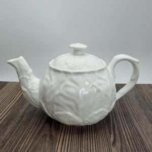 May include: A white ceramic teapot with a lid and handle. The teapot has a textured, leaf-like pattern covering its surface. The spout is on the left, and the handle is on the right. The teapot is sitting on a wooden surface.