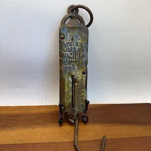 May include: An antique Excelsior Improved Spring Balance scale by Sargent & Co. USA. The scale is made of aged metal with a hook at the bottom and a ring at the top. The scale has a measurement gauge and is mounted on a black metal stand.