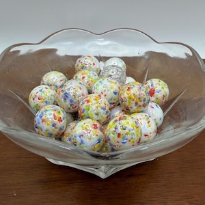 May include: A clear glass bowl filled with colorful marbles. The marbles are white with a speckled pattern of red, yellow, blue, and green. The bowl has a scalloped edge and sits on a wooden surface.