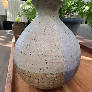 May include: A speckled ceramic vase with a wide body and a narrow neck. The vase features a color gradient, transitioning from a brown base to a light gray and blue upper section. The vase is displayed on a wooden tray.