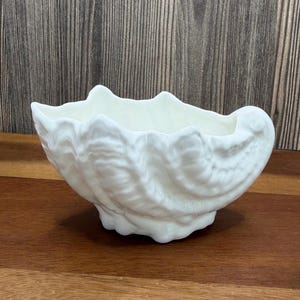 May include: A white ceramic seashell-shaped bowl with a textured surface. The bowl has a scalloped rim and sits on a small base. The bowl is displayed on a wooden surface.