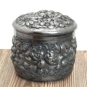 May include: A silver-toned, round trinket box with a lid. The box and lid are heavily embossed with floral designs, including detailed flowers and leaves. The box has a slightly aged appearance.
