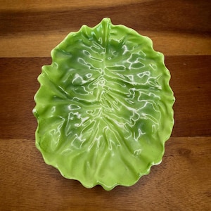 May include: A ceramic, leaf-shaped dish in a vibrant green color. The dish has a detailed, textured surface mimicking the veins and shape of a lettuce leaf. The edges are gently scalloped, adding to the naturalistic design. The dish is set against a wooden background.