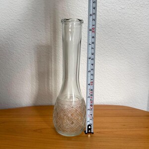 Pu&ograve; includere: Un vaso di vetro trasparente con un motivo intrecciato strutturato alla base. Il vaso &egrave; alto circa 20 cm. Il vaso ha un collo lungo e sottile e una base svasata. Il vaso &egrave; appoggiato su una superficie di legno.