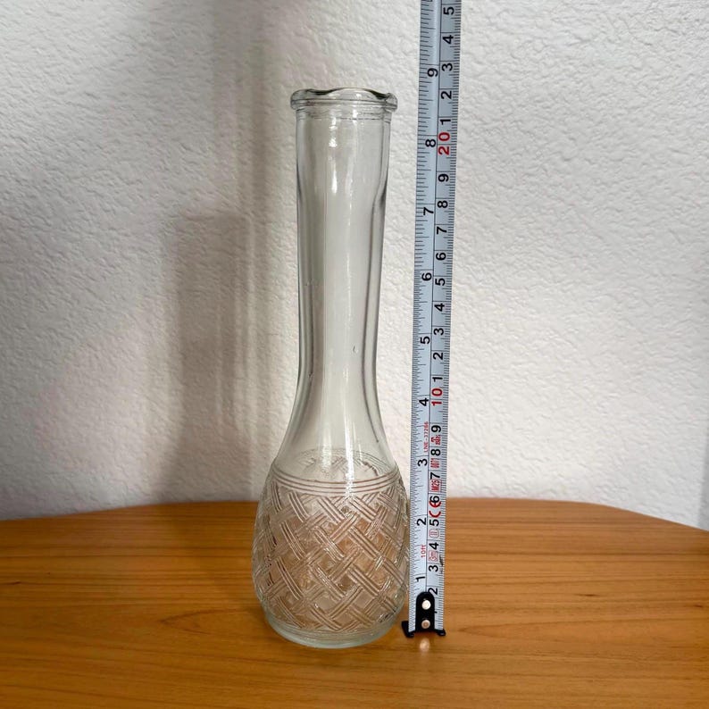 Pu&ograve; includere: Un vaso di vetro trasparente con un motivo intrecciato strutturato alla base. Il vaso &egrave; alto circa 20 cm. Il vaso ha un collo lungo e sottile e una base svasata. Il vaso &egrave; appoggiato su una superficie di legno.