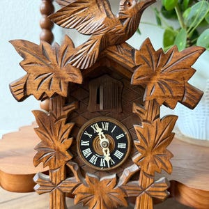 May include: A brown wooden cuckoo clock featuring intricate hand-carved maple leaf details and a bird perched on top. The clock face is circular with a black background, white Roman numerals, and ornate gold-colored hands. A small wooden door is visible.