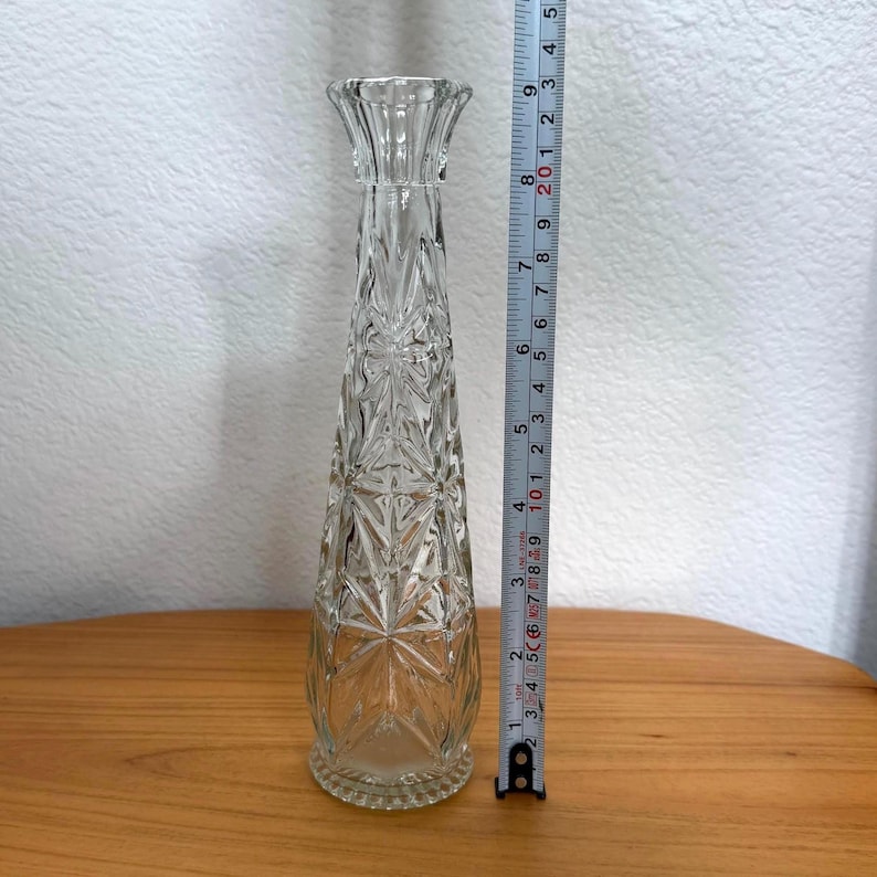 Pu&ograve; includere: Un vaso di vetro trasparente con un motivo a stella strutturato. Il vaso &egrave; alto circa 20 cm. Il vaso ha una parte superiore svasata e una base rotonda. Il vaso &egrave; appoggiato su una superficie di legno.