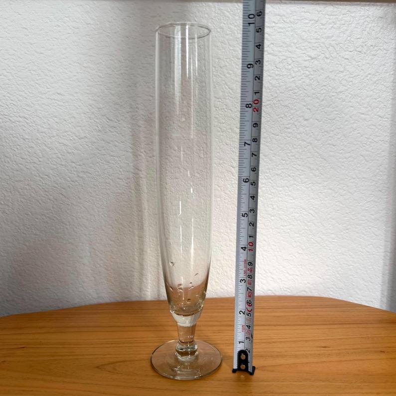 Pu&ograve; includere: Un vaso alto di vetro trasparente con un corpo sottile e cilindrico e una base svasata. Il vaso &egrave; alto circa 23 cm. &Egrave; appoggiato su una superficie di legno, con un metro a nastro accanto.