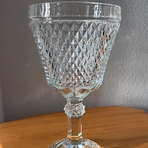 May include: A clear, vintage-style wine glass with a diamond-cut pattern on the bowl. The glass has a stemmed base and a wide rim. The glass is made of clear glass and sits on a wooden surface.