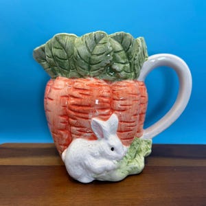 May include: A ceramic pitcher shaped like a carrot with green leaves and a white handle. A white bunny is nestled at the base of the carrot. The pitcher is orange, green, and white, perfect for Easter or spring decor.