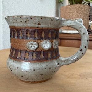 May include: Handmade ceramic mug with a speckled, light gray glaze. The mug features a brown band with vertical indentations and three circular, white, flower-like designs. The handle is curved and has a decorative element at the top.