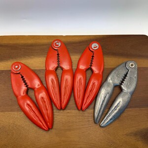 May include: Four crab crackers on a wooden surface. Three are bright red, and one is silver. The crackers are shaped like crab claws, with a hinged design for cracking shells. The red ones have a glossy finish.