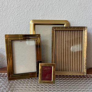May include: A collection of four rectangular picture frames in various sizes and gold-toned finishes. The smallest frame features a maroon insert with the text "Solid Brass 2" x 3"" printed in white. The frames have smooth and textured borders.