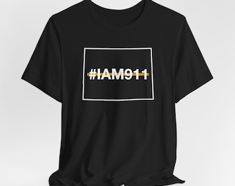 Op de afbeelding: Zwart T-shirt met de tekst "#IAM911" in het wit, omlijnd door een witte rechthoek. Een gouden horizontale lijn kruist de tekst. Het T-shirt is gemaakt van een zacht materiaal.