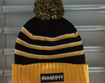 Op de afbeelding: Zwarte en gele gestreepte gebreide beanie met een gele pompon bovenop. De beanie heeft een gele boord met een zwart patch waarop "#IAM911" staat.