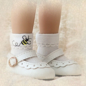 May include: White baby shoes with a buckle closure and a white lace trim. The shoes are worn by a doll with white socks that have a bee embroidered on them.