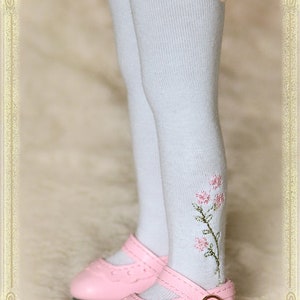 May include: White knee-high socks with a delicate lace trim and embroidered pink flowers. The socks are paired with pink leather shoes with a buckle.