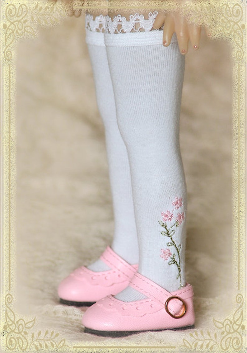 May include: White knee-high socks with a delicate lace trim and embroidered pink flowers. The socks are paired with pink leather shoes with a buckle.