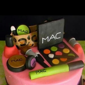 May include: A pink cake decorated with makeup themed fondant. The cake features a makeup palette, lipstick, nail polish, and a makeup bag with the word "MAC" on it.