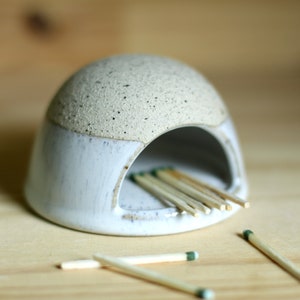 May include: A white ceramic match holder with a speckled glaze. The holder has a rounded top and a small opening for matches. Matches are inside the holder and some are scattered on a wooden surface.