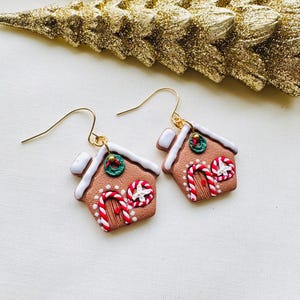 May include: A pair of gingerbread house earrings with gold-colored hooks. The earrings are brown with white icing accents, candy cane details, and a small wreath. The background includes a gold glittery Christmas tree.