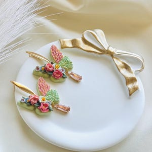 May include: A pair of delicate butterfly earrings with floral accents. The earrings are made of polymer clay and feature a mix of pink, green, and white flowers. Each earring has a small, teardrop-shaped crystal dangling from the bottom.