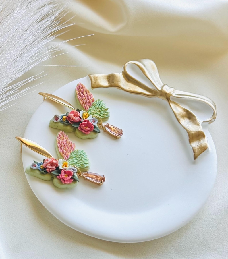 May include: A pair of delicate butterfly earrings with floral accents. The earrings are made of polymer clay and feature a mix of pink, green, and white flowers. Each earring has a small, teardrop-shaped crystal dangling from the bottom.