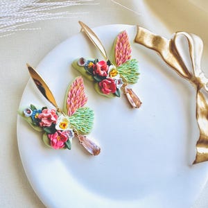 May include: A pair of gold butterfly earrings with pink, green, and white floral accents. The earrings have a teardrop-shaped pink crystal dangling from each butterfly.