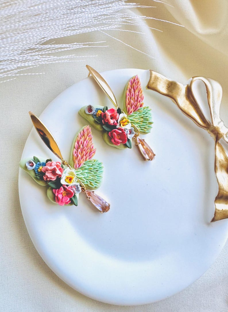 May include: A pair of gold butterfly earrings with pink, green, and white floral accents. The earrings have a teardrop-shaped pink crystal dangling from each butterfly.